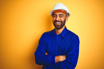 Handsome indian worker man wearing uniform and helmet over isolated yellow background happy face smiling with crossed arms looking at the camera. Positive person.