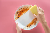 Fototapeta Tęcza - woman using yellow cleaning sponge to clean up and washing food stains and dirt on white dish after eating meal isolated on pink background. cleaning , healthcare and sanitation at home concept