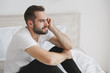 © ViDi Studio - Calm young handsome bearded man sitting in bed with white sheet pillow blanket in bedroom at home. Sad upset male spending time in room. Rest relax good mood lifestyle concept. Mock up copy space.