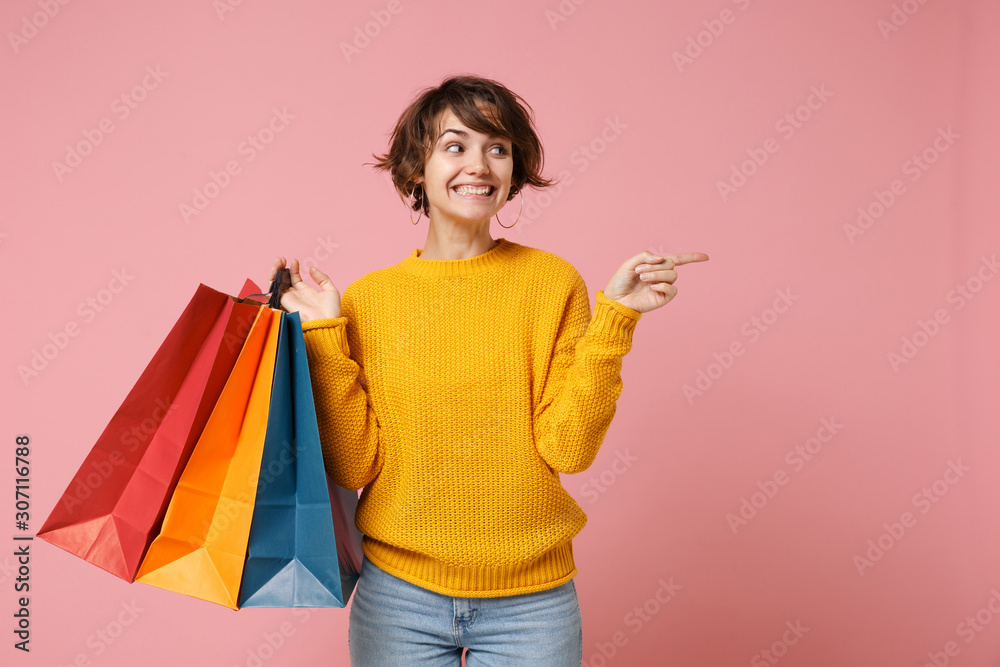 Smiling young brunette woman girl in yellow sweater posing isolated on pink background. People lifestyle concept. Mock up copy space. Hold package bag with purchases after shopping point finger aside. - obrazy, fototapety, plakaty 