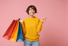 Smiling Young Brunette Woman Girl In Yellow Sweater Posing Isolated On Pink Background. People Lifestyle Concept. Mock Up Copy Space. Hold Package Bag With Purchases After Shopping Point Finger Aside.