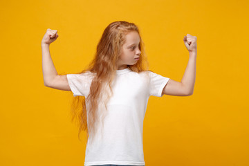 Wall Mural - Strong little ginger kid girl 12-13 years old in white t-shirt isolated on bright yellow background children studio portrait. Childhood lifestyle concept. Mock up copy space. Showing biceps muscles.