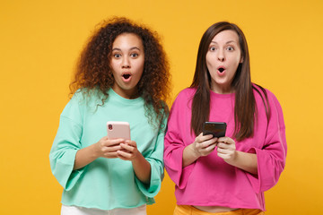 Sticker - Two shocked women friends european african american girls in pink green clothes posing isolated on yellow background. People lifestyle concept. Mock up copy space. Using mobile phone, typing message.