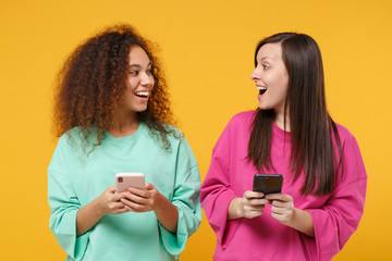 Sticker - Two excited women friends european african american girls in pink green clothes posing isolated on yellow background. People lifestyle concept. Mock up copy space. Using mobile phone, typing message.