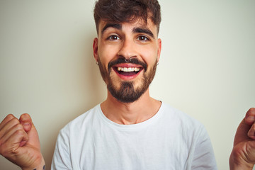 Young man with tattoo wearing t-shirt standing over isolated white background celebrating surprised and amazed for success with arms raised and open eyes. Winner concept.