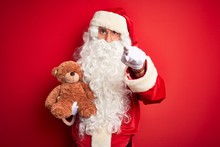 Middle Age Man Wearing Santa Claus Costume Holding Teddy Bear Over Isolated Red Background Pointing With Finger To The Camera And To You, Hand Sign, Positive And Confident Gesture From The Front
