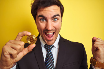 Wall Mural - Young handsome businessman holding bitcoin standing over isolated yellow background screaming proud and celebrating victory and success very excited, cheering emotion