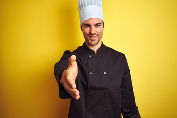 Wall Mural - Young chef man wearing uniform and hat standing over isolated yellow background smiling friendly offering handshake as greeting and welcoming. Successful business.