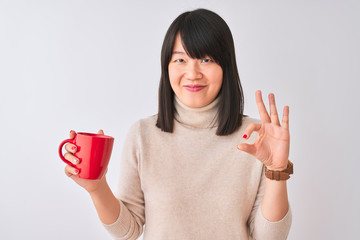 Wall Mural - Young beautiful Chinese woman drinking red cup of coffee over isolated white background doing ok sign with fingers, excellent symbol