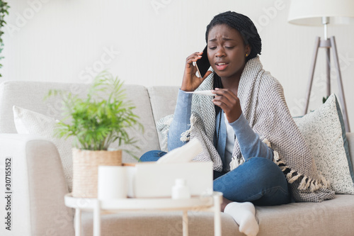 Cuadros en Lienzo Sick woman sitting on sofa, holding thermometer and calling family doctor