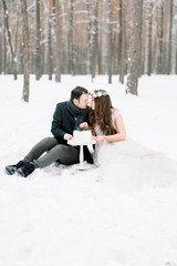  A couple in a wedding clothes sitting and cutting their wedding cake, against the background of the winter forest. Romantic date