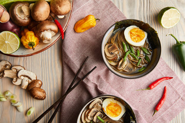 top view of traditional spicy ramen in bowls with chopsticks and vegetables on wooden table with napkin