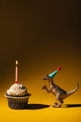 Wall Mural - Toy dinosaur in party cap beside cupcake with burning candle on brown background