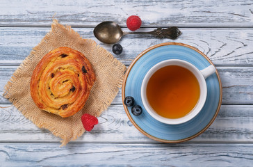 Sweet puff bun with cream and raisins in the shape of a snail on burlap. Tasty delicious dessert, silver old teaspoon, cup of tea, berries of fresh juicy raspberries and blueberries. French breakfast.