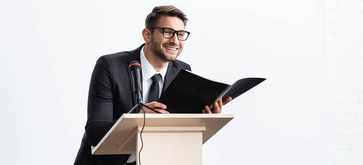 Wall Mural - panoramic shot of smiling businessman in suit standing at podium tribune and holding folder during conference isolated on white