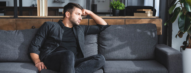 Wall Mural - panoramic shot of handsome and pensive man sitting on sofa in apartment