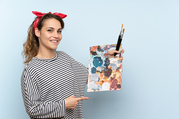 Wall Mural - Young artist woman holding a palette over isolated blue background pointing to the side to present a product