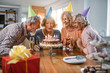 © Drazen - Senior birthday woman blowing candles on a cake while celebrating with friends at home.