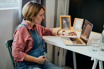 Attractive caucasian pregnant woman sitting at home office, touching belly and using laptop for work.