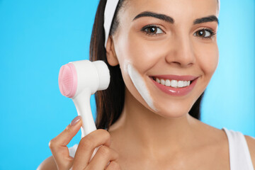 Sticker - Young woman using facial cleansing brush on light blue background, closeup. Washing accessory