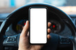 Close-up of male hand holding smartphone with white mockup on screen, background of car steering wheel.