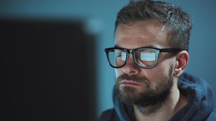 Bearded male hacker in hoodie and glasses working on a computer in a dark office room. Cybercrime concept
