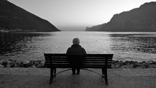 A Lonely Old Man Sits On A Wooden Bench At Dusk After Sunset, Looking At The Lake And The Light On The Horizon.  