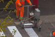 © Shcherbyna - Traffic line painting. Workers are painting white street lines on pedestrian crossing. Road cones with orange and white stripes in background, standing on asphalt during road construction works