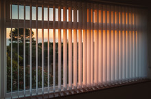White Vertical Slat Blinds Hanging In Front Of A Window As The Sun Is Setting Turning The Light Golden. The Slats Have Sealed Glued Pockets And No Cords At The Bottom.