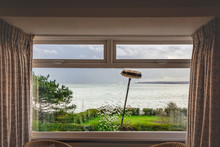 A Double Glazed Picture Window With Small Top Light Opening Windows Being Cleaned With An Extension Brush And Pure Water. Some Motion Blur Showing The Movement Of The Brush. There Is A Sea View.