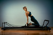 © Georgii - Young girl doing pilates exercises with a reformer bed. Beautiful slim fitness trainer on reformer gray background, low key, art light. Fitness concept