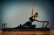 © Georgii - Young girl doing pilates exercises with a reformer bed. Beautiful slim fitness trainer on reformer gray background, low key, art light. Fitness concept