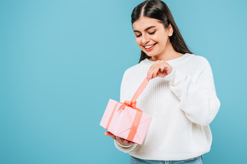 smiling pretty girl in white sweater unwrapping gift box isolated on blue