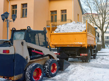 Small Loader Removing And Loading Snow Into A Truck. Skid Steer Loading Snow On Dump Truck. Cleaning City Street After Blizzard Or Snowfall. Wheel Loader Clearing And Removing Snow In Winter