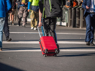 Traveller carrying backpack and small red suitcase walking alone on busy street.