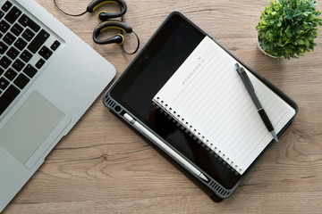 Wood office desk table with blank notebook  over tablet computer and notebook, pen, earphone. Top view.