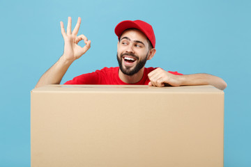 Wall Mural - Delivery man in red uniform isolated on blue background, studio portrait. Male employee in cap t-shirt print working as courier dealer hold empty cardboard box. Service concept. Mock up copy space.