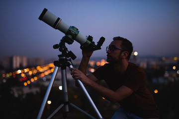 Wall Mural - astronomer with a telescope watching at the stars and moon with blurred city lights in the backgroun