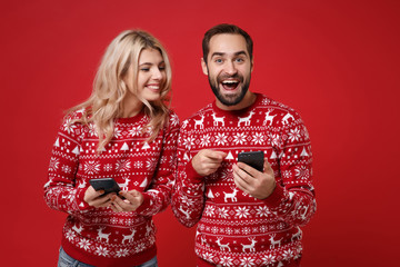 Wall Mural - Excited young couple guy girl in Christmas knitted sweaters posing isolated on red background. New Year 2020 celebration holiday concept. Mock up copy space. Using mobile phone typing sms message.