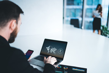 Executive man in suit sitting at desk in office typing on laptop while browsing smartphone in workspace in company