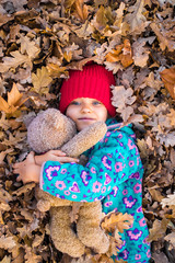  Little young girl holds teddy bear toy, lays in yellow leaves heap. Outdoor forest location, sunny day, nature.