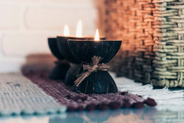 Wall Mural - Burning spa aroma candle in coconut shell on a knitted multicolored rug and wicker box with a towel opposite the white wall, cozy home interior.