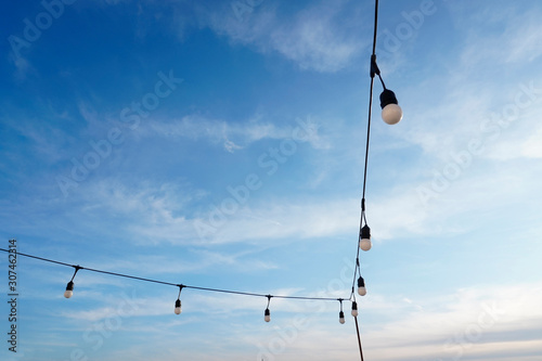 Cuadros en Lienzo garland of large bulbs on the terrace for decoration on holidays or at night against a blue sky background with small white clouds