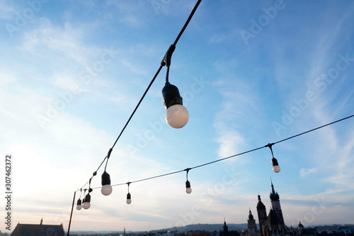 Fotomural garland of large bulbs on the terrace for decoration on holidays or at night against a blue sky and old buildings background with small white clouds