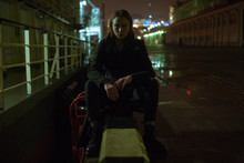 Girl In Black Coat Sitting And Posing On Pier At Night