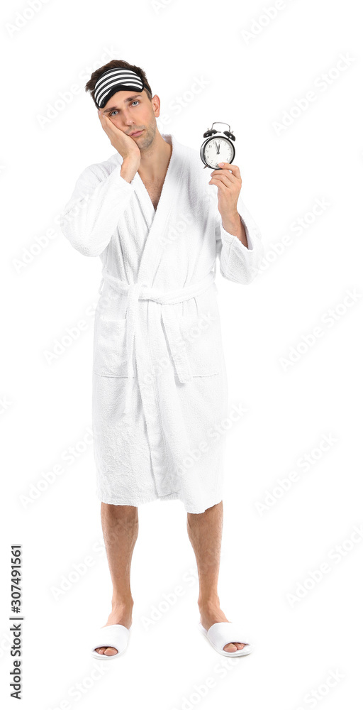 Tired man in bathrobe, sleep mask and alarm clock on white background