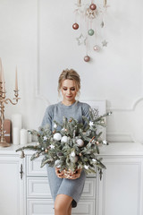 Wall Mural - Fashionable young leggy woman with platinum blond hair in knitted dress posing with Christmas decorations. Model girl with evening makeup and hairstyle posing in beautiful New year interior