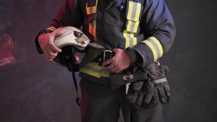 Wall Mural - Close-up view of a brave firefighter in special uniform holds protective helmet standing against a gray wall