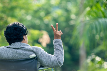 Asia elderly woman sitting on wheelchair and shows two fingers outdoor,Trees in the summer background.Have a nice day.