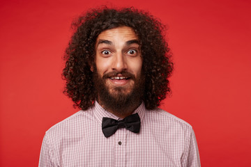 Surprised pretty young brunette male with beard rounding his brown eyes while looking to camera, raising eyebrows amazedly and wrinkling forehead while posing against red background